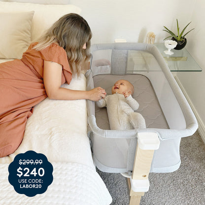 A woman wearing a brown dress lies on a bed, holding hands with a smiling baby resting in the Newton Baby Bassinet & Bedside Sleeper in gray. The price tag displays $299.99 crossed out, now $240 with code LABOR20. A glass shelf decorated with decor is visible in the background.