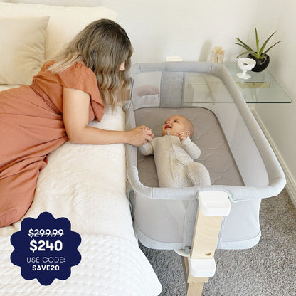 A woman in a rust-colored dress lies on a bed, smiling and holding hands with her baby resting in the Newton Baby Bassinet & Bedside Sleeper. A promo label shows $299.99 $240 USE CODE: SAVE20 in the lower left corner.