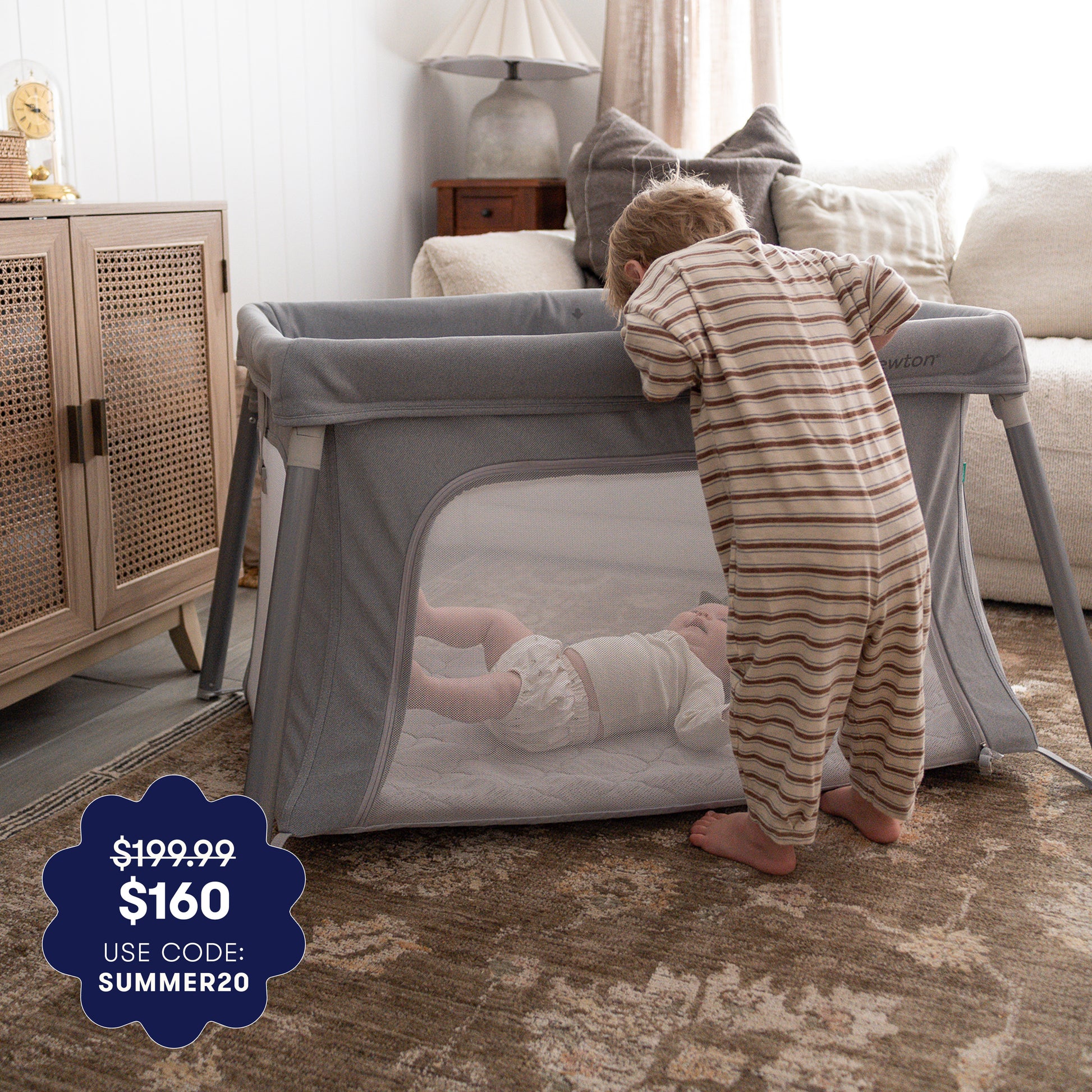 A toddler in striped pajamas peers into the Newton Baby Compact Travel Crib & Play Yard, where a baby rests on an ultra-breathable mattress. A graphic shows $199.99 slashed to $160 with code SUMMER20. The scene is set in a cozy, carpeted living room. 999-5101-GRY 