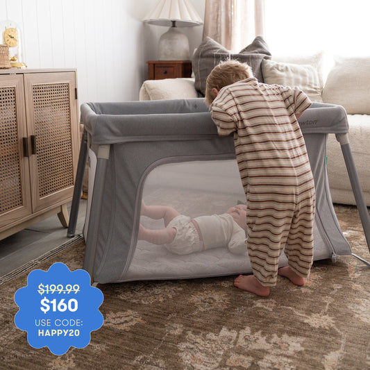 A toddler in striped pajamas peers into a Newton Baby Compact Travel Crib & Play Yard with a breathable mattress, where a baby lies. Price: $199.99 $160 with code HAPPY20. The cozy living room is decorated in neutral tones.