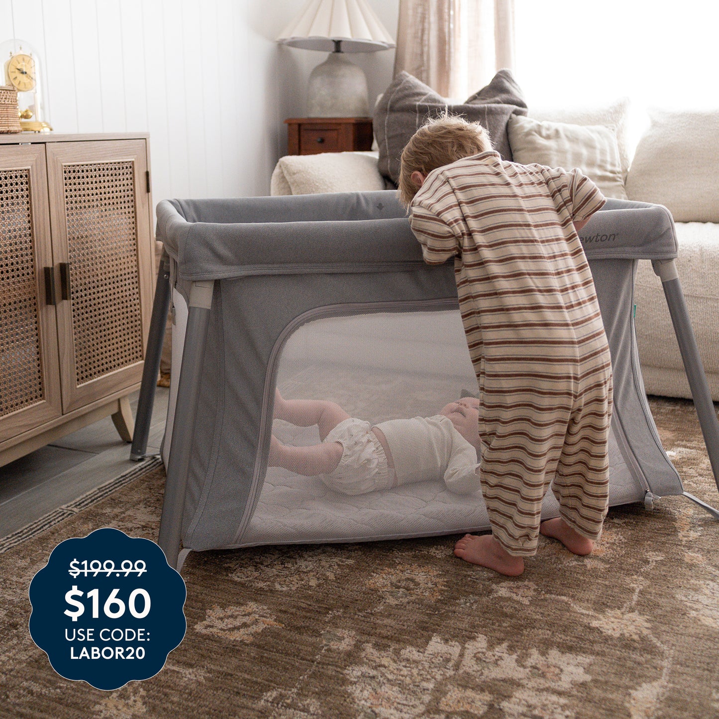 A toddler in striped pajamas looks into a gray Newton Baby Compact Travel Crib & Play Yard, where a baby is inside resting on its breathable mattress for added comfort. A price tag in the corner reads $199.99 $160 USE CODE: LABOR20. The room features a rug, lamp, and wooden furniture. 999-5101-GRY