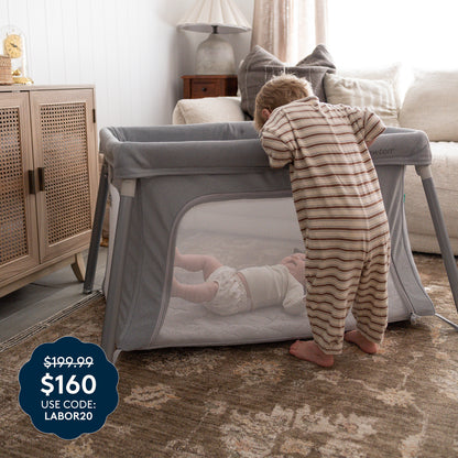 A toddler in striped pajamas looks into a gray Newton Baby Compact Travel Crib & Play Yard, where a baby is inside resting on its breathable mattress for added comfort. A price tag in the corner reads $199.99 $160 USE CODE: LABOR20. The room features a rug, lamp, and wooden furniture. 999-5101-GRY