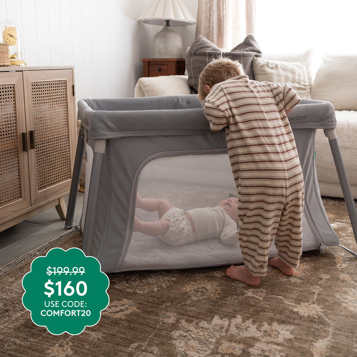 A toddler gazes into a gray Compact Travel Crib & Play Yard by Newton Baby, where a baby rests on a breathable mattress. Price: $199.99, now $160 with code COMFORT20. A rug, cabinet, and lamp are in the background.