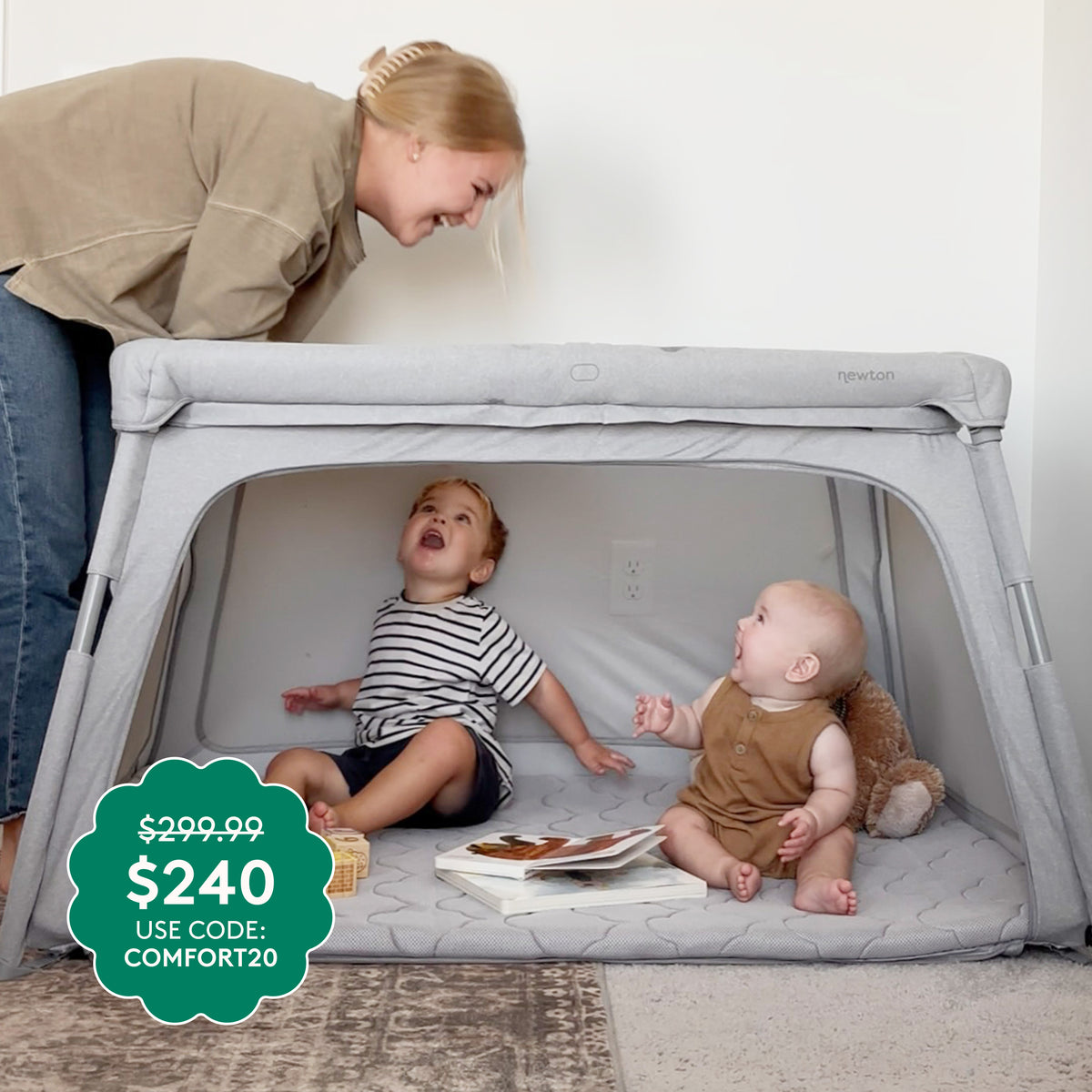 A woman smiles at two toddlers in a Deluxe Travel Crib & Play Yard by Newton Baby. Teddy bear and book inside. Green sticker: $299.99 drops to $240 with code COMFORT20.