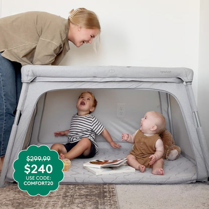A woman smiles at two toddlers in a Deluxe Travel Crib & Play Yard by Newton Baby. Teddy bear and book inside. Green sticker: $299.99 drops to $240 with code COMFORT20.