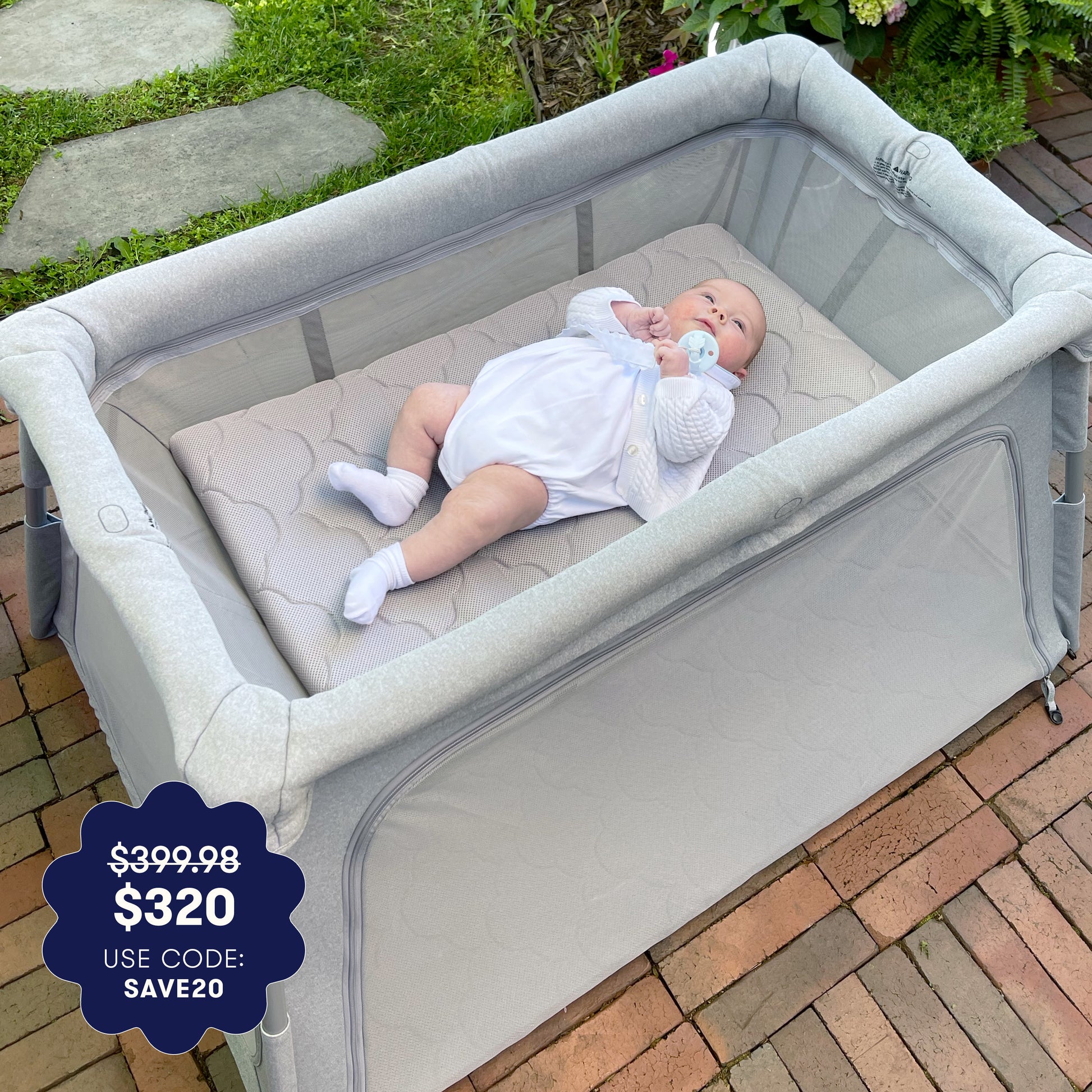 A baby in a white outfit rests on a washable mattress inside the Newton Baby Deluxe Travel Crib & Play Yard with gray mesh, on a brick patio. Price dropped from $399.98 to $320—use code SAVE20 for an extra discount. 