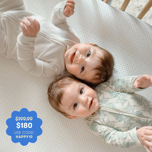 Two smiling babies in zip-up outfits lie on the Newton Baby Essential Crib Mattress. A blue tag shows: "$199.99 $180 USE CODE: HAPPY10.