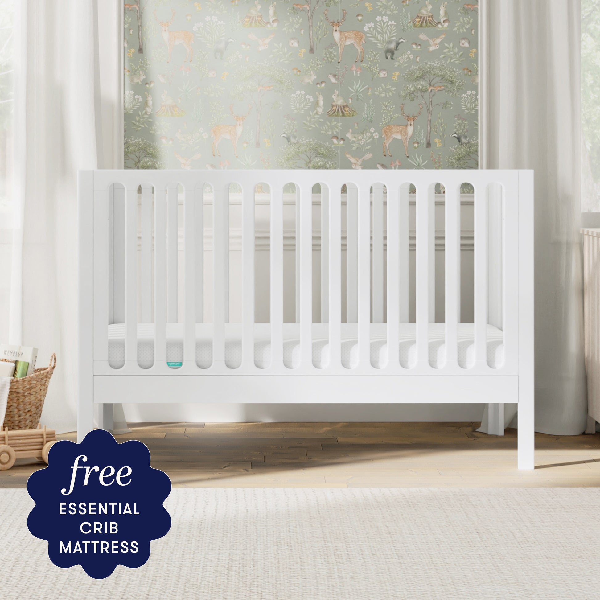 A white Newton Galileo Convertible Crib by Newton Baby, complete with a mattress, stands in a bright nursery decorated with nature-themed wallpaper. A badge in the corner reads: “Free ESSENTIAL CRIB MATTRESS.”.