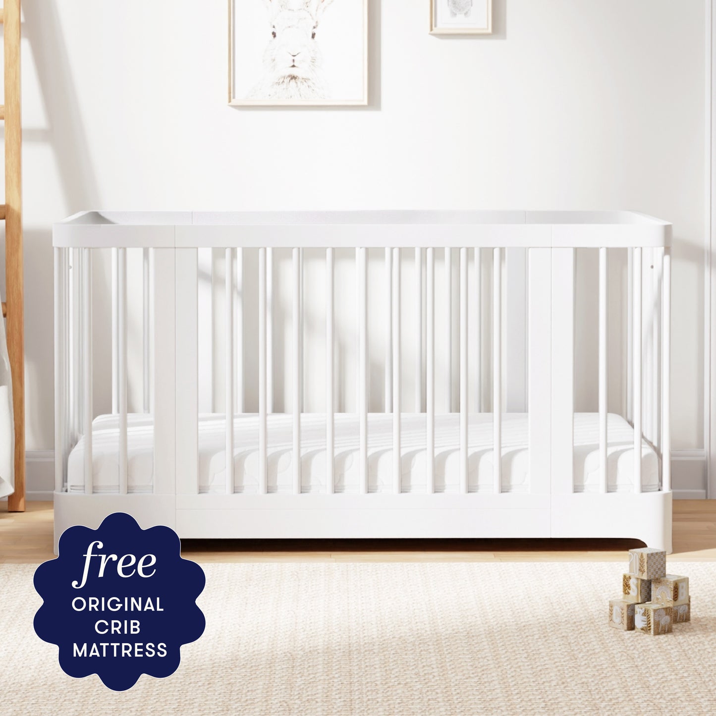 The Newton Nest Convertible Crib by Newton Baby, featuring modern white vertical slats, stands on a light carpet in the nursery. A sign reads “free original crib mattress.” Stacked wooden blocks are placed near the crib.