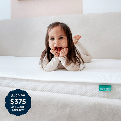 A young child smiles and lounges on the Kids’ Twin Mattress by Newton Baby, which features a white, washable cover and a beige headboard. A sale badge in the corner displays $375, was $499.99, use code: LABOR25. The breathable mattress is GREENGUARD Gold certified for your peace of mind and includes the signature green Newton tag.