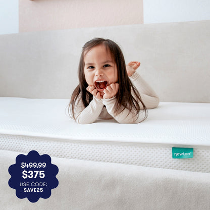 A young child with long brown hair smiles on a white, breathable Newton Baby Kids' Twin Mattress. Price tag: $499.99 $375 USE CODE: SAVE25. The mattress label reads "newton.