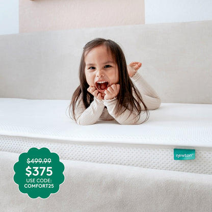 A smiling child lies on a white, GREENGUARD Gold certified Kids’ Twin Mattress by Newton Baby in a bright room. Green price tag: $499.99 $375 USE CODE: COMFORT25. Small blue newton label in the corner.