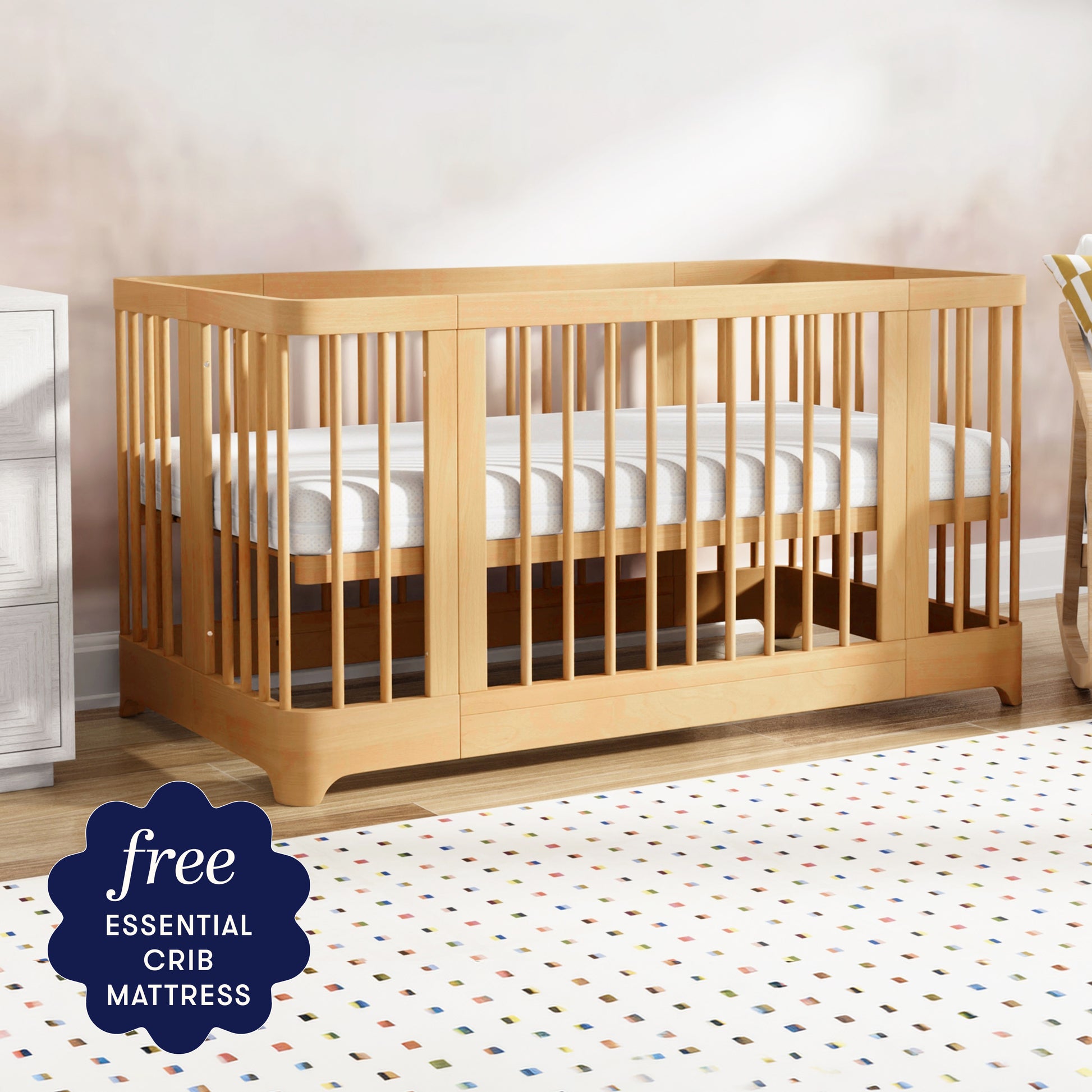The Newton Nest Convertible Crib by Newton Baby, shown here in natural wood with a white mattress, brightens up the nursery atop a colorful dotted rug. A corner sticker announces: free ESSENTIAL CRIB MATTRESS. GREENGUARD Gold certified, this crib ensures your baby’s sleep space is both stylish and healthy.
