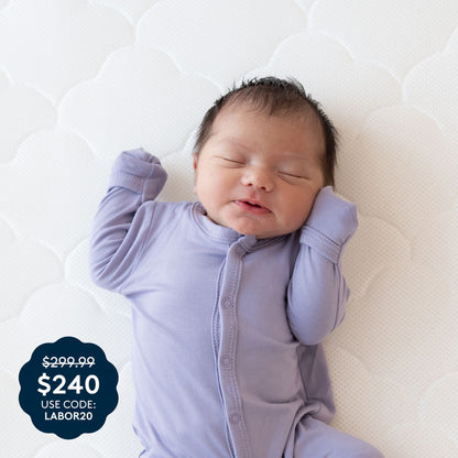 A newborn baby in a lavender onesie smiles with eyes closed while lying on a white textured surface atop a breathable Newton Baby Crib Mattress. A promo graphic features: $299.99 $240 USE CODE: LABOR20, displayed in white text on a dark scalloped background.