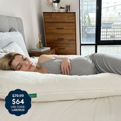A woman in gray pajamas sleeps on The Pregnancy Pillow by Newton Baby, a large white U-shaped pillow designed for ultimate comfort and support, in a bright bedroom. The price tag reads $79.99, now $64 with code LABOR20.
