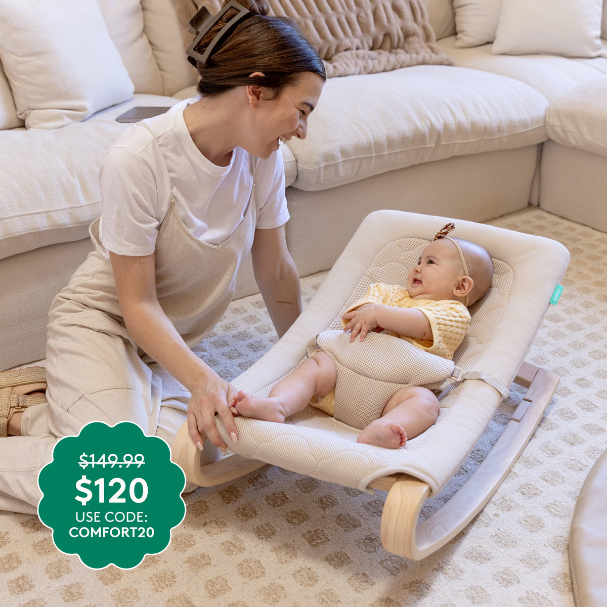 A woman smiles at a baby in a Newton Baby Rocker by Newton Baby on a patterned beige carpet. A discount tag shows $149.99 $120 USE CODE: COMFORT20, with a white couch in the background.