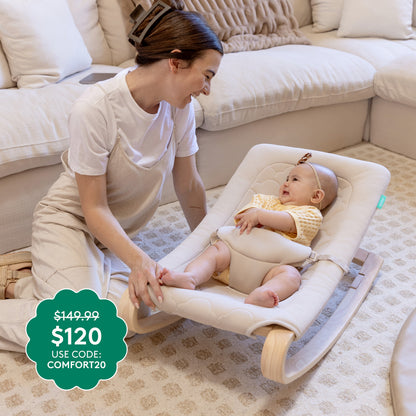 A woman smiles at a baby in a Newton Baby Rocker by Newton Baby on a patterned beige carpet. A discount tag shows $149.99 $120 USE CODE: COMFORT20, with a white couch in the background.