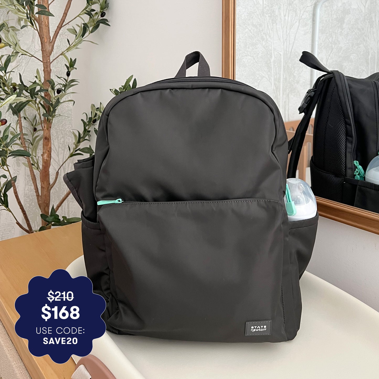 A stylish Newton X STATE Lorimer Diaper Bag by Newton Baby in black with mint zippers sits on a table near a plant and mirror. A blue badge shows the price drop from $210 to $168 with code SAVE20. A baby bottle is in the side pocket.