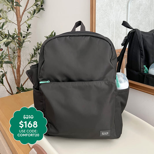 A stylish Newton X STATE Lorimer Diaper Bag with teal zippers rests near a plant and mirror. Price: $210 $168—use code: COMFORT20. A small container peeks from the side pocket.