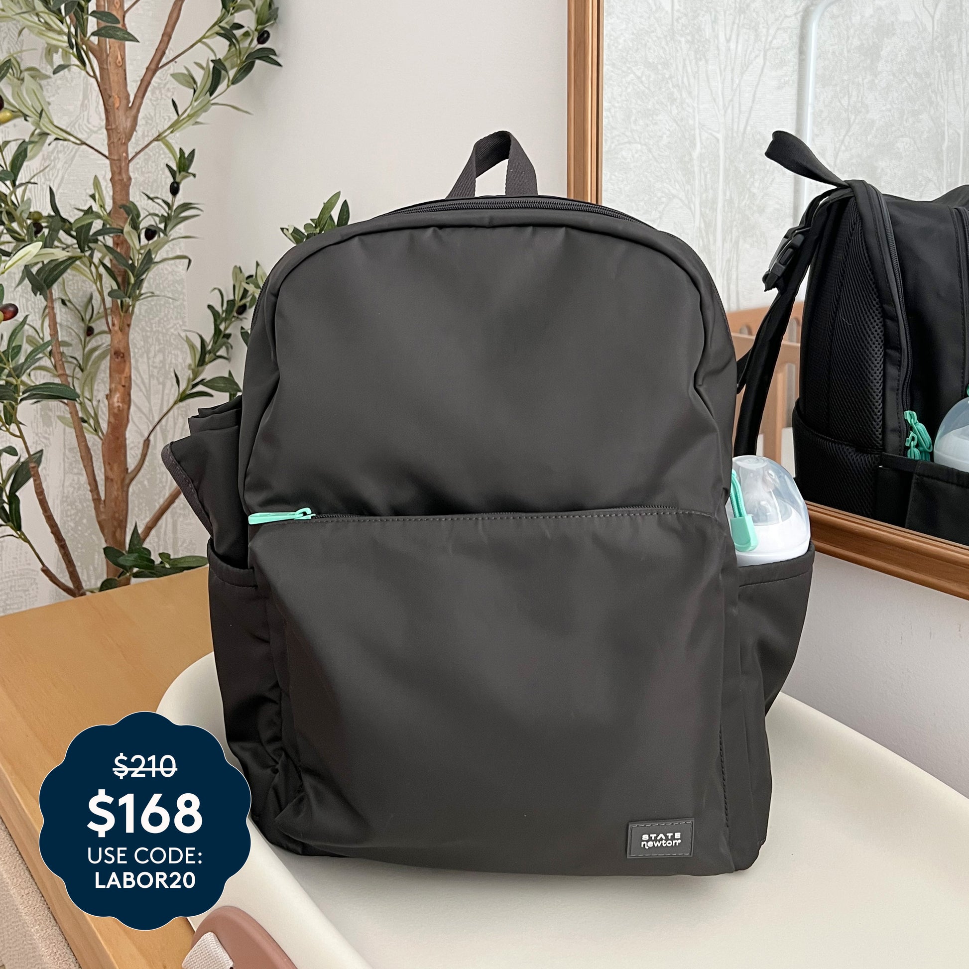 The Newton Baby x STATE Lorimer Diaper Bag rests stylishly on a beige surface, accompanied by a mirror and a leafy plant. This fashionable diaper backpack features a breathable mesh back panel for extra comfort. A blue price tag displays $168 with code LABOR20, marked down from $210. A small container peeks out from the side pocket.