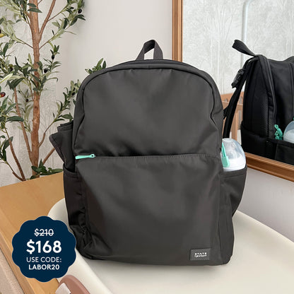 The Newton Baby x STATE Lorimer Diaper Bag rests stylishly on a beige surface, accompanied by a mirror and a leafy plant. This fashionable diaper backpack features a breathable mesh back panel for extra comfort. A blue price tag displays $168 with code LABOR20, marked down from $210. A small container peeks out from the side pocket.