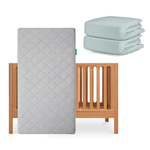 The Newton Baby Galileo Crib with the Original Crib Mattress stands upright, while two folded pale blue organic cotton sheets are stacked neatly beside it. skip