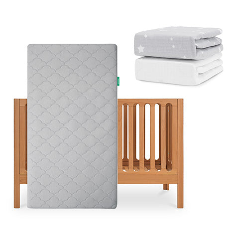 The Newton Baby Galileo Crib stands upright, paired with the Original Crib Mattress in a gray pattern. Behind it are two folded fitted sheets—one organic white cotton and one gray with stars—from the Newton Baby collection. skip