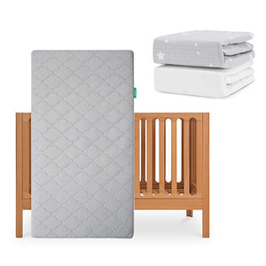 The Newton Baby Galileo Crib stands upright, paired with the Original Crib Mattress in a gray pattern. Behind it are two folded fitted sheets—one organic white cotton and one gray with stars—from the Newton Baby collection. skip