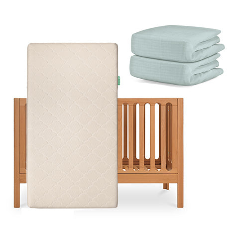 A Newton Baby Galileo Crib with an Original Crib Mattress standing in front, plus two folded light blue organic cotton sheets stacked beside the crib. skip