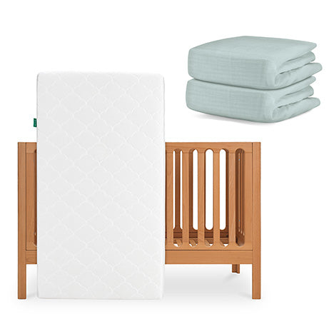 The Newton Baby Galileo Crib with the Original Crib Mattress is shown with two folded light blue organic cotton sheets stacked beside it. skip