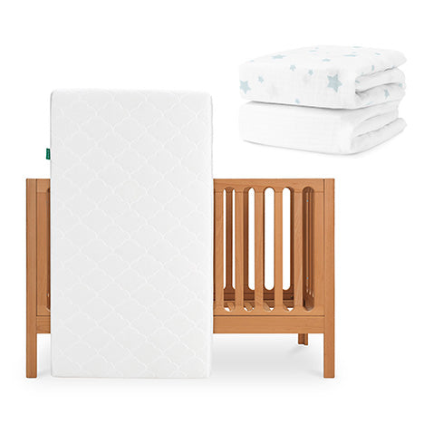 The Newton Baby Galileo Crib is shown with the Original Crib Mattress propped against it. In the upper right, two folded white Newton Baby sheets are visible—one featuring a blue star pattern. skip