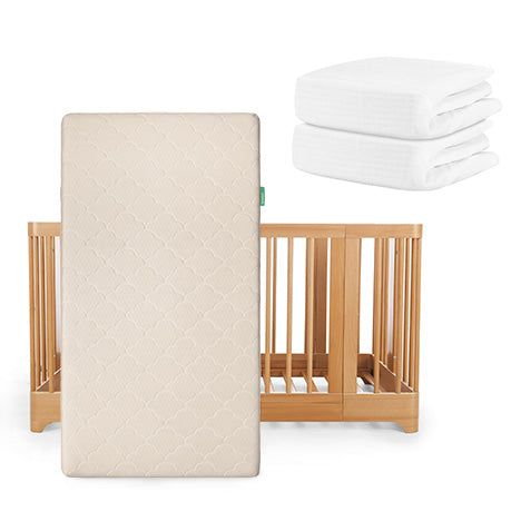The Newton Baby Nest Convertible Crib is paired with the Original Mattress in beige (propped upright) and two folded white sheets from the Nest Sleep Bundle stacked nearby.
