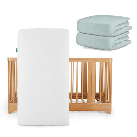 A Newton Baby Nest Convertible Crib with the Original Mattress stands upright, while two folded Breathable Organic Cotton Sheets in light blue rest nearby for added comfort.