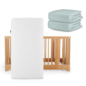 A Newton Baby Nest Convertible Crib with the Original Mattress stands upright, while two folded Breathable Organic Cotton Sheets in light blue rest nearby for added comfort.
