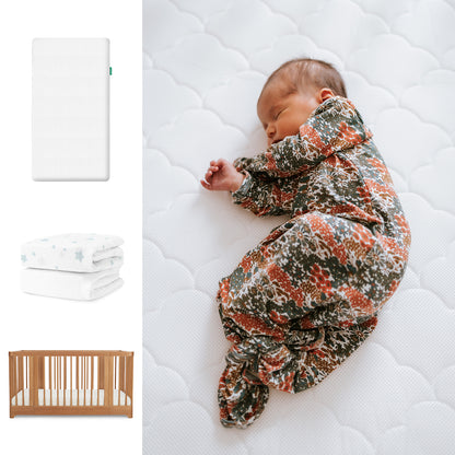 A baby in a floral swaddle sleeps on the Newton Baby Nest Convertible Crib with the Original Mattress and organic cotton sheets, next to a folded star-patterned blanket. 111-8420-CST-1000-WHT-SDW