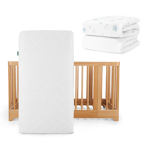 The Newton Baby Nest Convertible Crib with Original Mattress stands upright, topped with a folded white sheet featuring blue stars, while a Breathable Organic Cotton Sheet from Newton Baby rests nearby in the background.