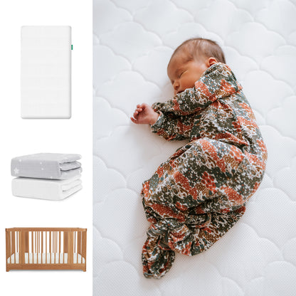 A newborn sleeps on the Newton Baby Nest Convertible Crib with the Original Mattress and organic cotton sheets, wearing a floral sleeper. Nearby are folded sheets and product images. 111-8420-CST-1000-WHT-SGW