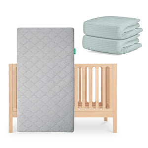 The Newton Baby Galileo Crib with the Original Crib Mattress in a subtle gray pattern stands nearby, while two folded light green organic cotton sheets are stacked in the background. skip