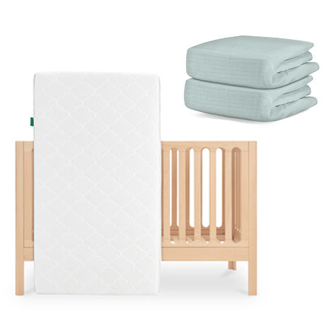 The Newton Baby Galileo Crib is shown with the Original Crib Mattress propped against it and two folded Newton Baby cotton sheets stacked neatly beside the crib. skip