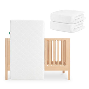 The Newton Baby Galileo Crib in light wood is paired with the Original Crib Mattress propped against it and two stacks of organic cotton sheets, all arranged on a plain white background.
skip