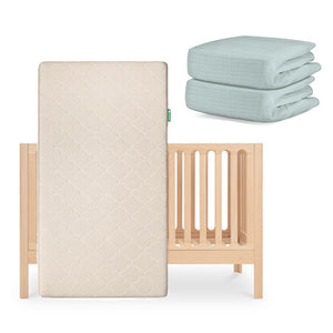 The Newton Baby Galileo Crib with the Original Crib Mattress stands upright, while two folded light blue organic cotton sheets are stacked neatly beside it. skip