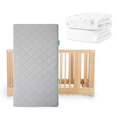 The Newton Baby Nest Convertible Crib with the Original Mattress is shown in light wood, paired with fitted sheets—one white with blue stars and one plain white—stacked to the right, all set against a clean white background.