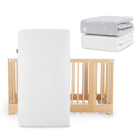 Up front is a Nest Convertible Crib with a white Original Mattress by Newton Baby. Behind, two breathable organic cotton crib sheets—one gray with white stars and one solid white—are neatly stacked.