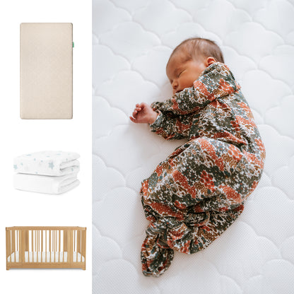 A baby in a floral sleeper rests on Newton Baby’s Nest Convertible Crib with the Original Mattress & Sheets. To the left are images of the beige mattress, a folded white sheet with blue stars, and the stylish crib. 111-8420-NAT-1000-SAN-SDW