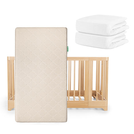 A Newton Baby Nest Convertible Crib with an Original Mattress stands upright, while two folded breathable organic cotton sheets from the same set are neatly stacked beside it.