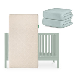 A light green Newton Baby Galileo Crib with the Original Crib Mattress propped upright in front and a stack of two breathable organic cotton sheets folded at the side. skip