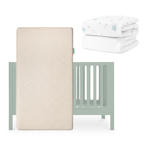 A light green Newton Baby Galileo Crib with an Original Crib Mattress stands in front, while two white crib sheets with blue stars—including a Breathable Organic Cotton Sheet—are stacked in the background. skip