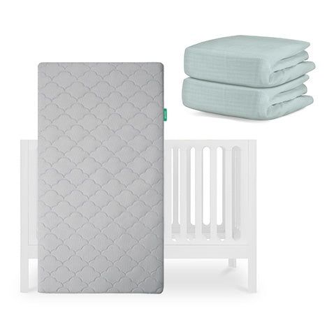 A Newton Baby Galileo Crib, Original Crib Mattress in gray stands upright before a white crib, with two light blue Newton Baby Breathable Organic Cotton Sheets folded neatly to the right. skip