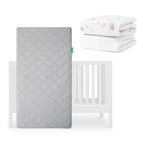 The Newton Baby Galileo Crib stands behind the Original Crib Mattress in gray, with organic cotton sheets and a white blanket with pink stars stacked nearby. skip