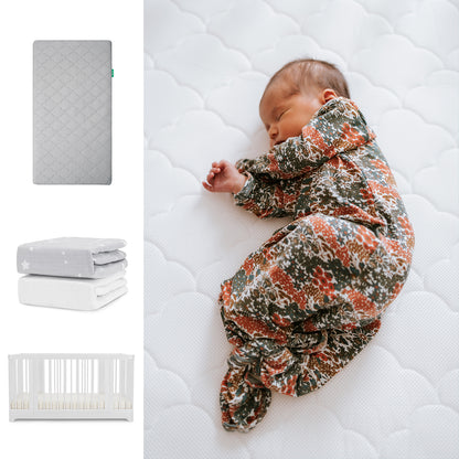 A newborn sleeps on Newton Baby's Nest Convertible Crib with the Original Mattress, wearing a floral swaddle. To the left are two folded Breathable Organic Cotton Sheets (gray and white) by Newton Baby. 111-8420-WHT-1000-GRY-SGW